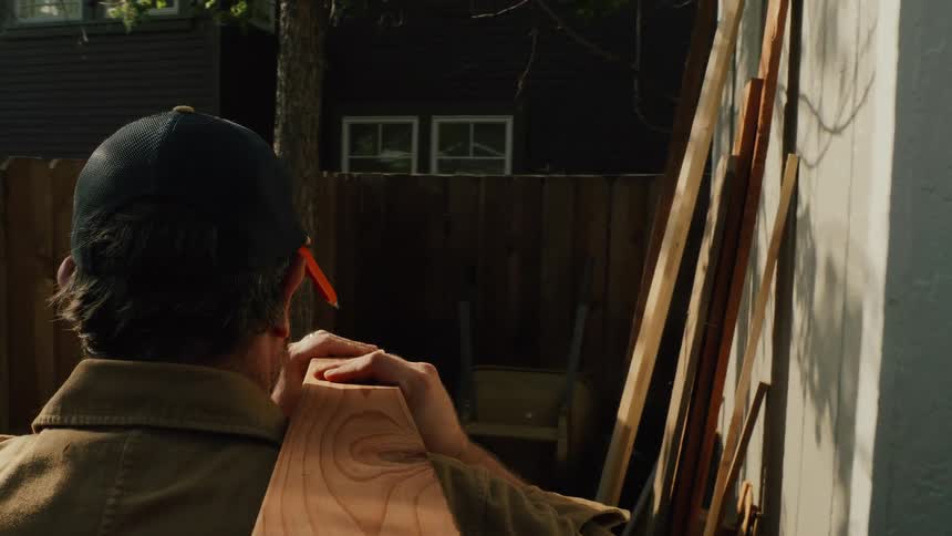 A handyman carries a large wooden plank on his shoulder through a sunny backyard in slow motion