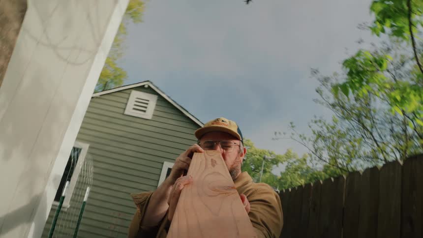 A low-angle shot follows a bearded carpenter as he picks up a wooden plank and sights down its length to check for straightness in his backyard