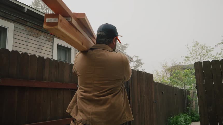 A carpenter wearing a tool belt and carrying wooden planks on his shoulder walks down a path in slow motion