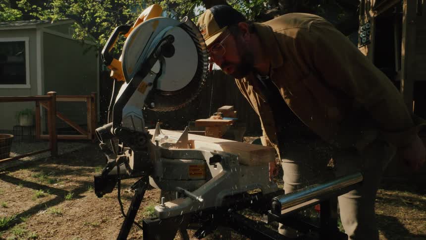 A man in his backyard carefully uses a miter saw to cut a piece of wood in slow motion, sending sawdust flying through the air