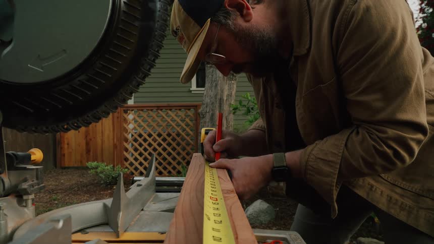 A focused woodworker carefully measures a wooden board with a tape measure and marks it with a pencil