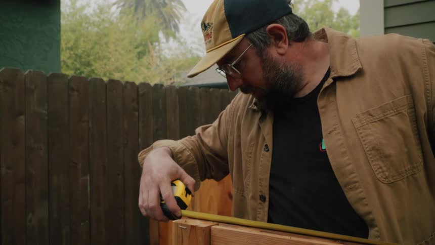 A handyman in a hat and glasses carefully uses a tape measure on a wooden project in his backyard