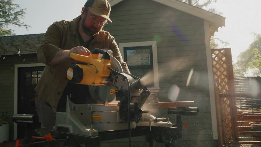 A man cuts a wooden plank with a miter saw in his backyard, sending sawdust flying through the sunny air in slow motion