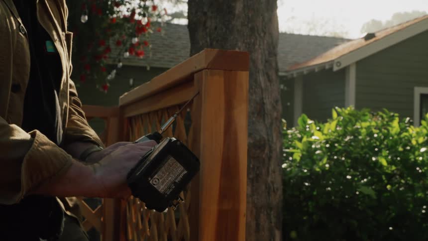 Slow motion shot of a bearded man in a hat carefully drilling a screw into a new wooden fence in a sunlit backyard