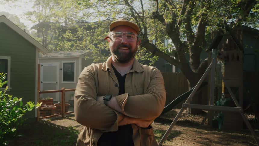 A confident, smiling man with a beard and glasses poses for a portrait in his sunny suburban backyard