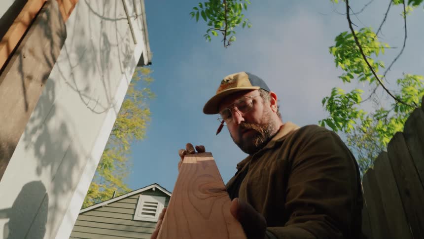 A low-angle shot reveals a focused carpenter marking a piece of lumber in his backyard on a sunny day