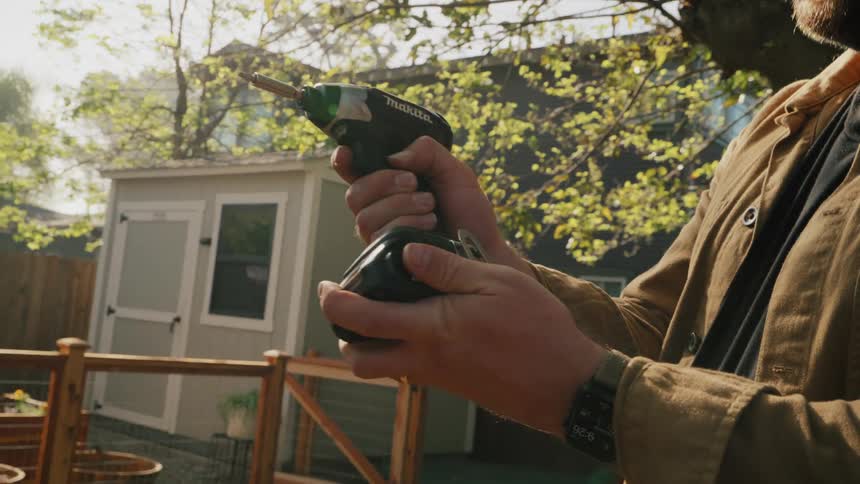 A man holds a power drill in his backyard as the camera slowly racks focus to the shed in the background