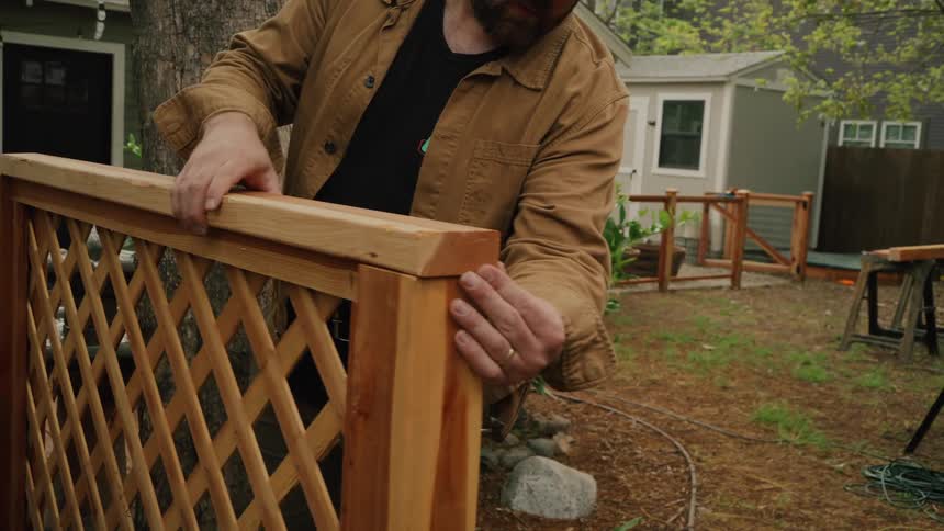 A skilled craftsman carefully inspects the newly installed top rail of a wooden lattice fence in his backyard
