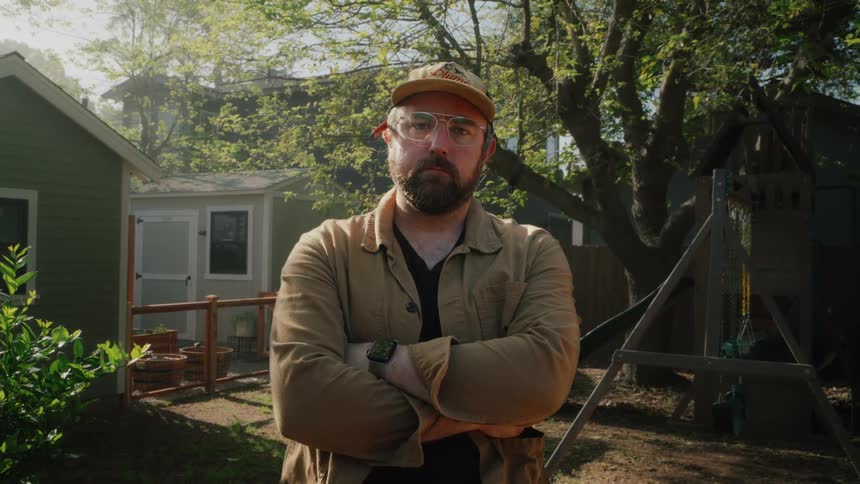 Portrait of a serious bearded man with glasses and a hat standing with his arms crossed in a sunny backyard