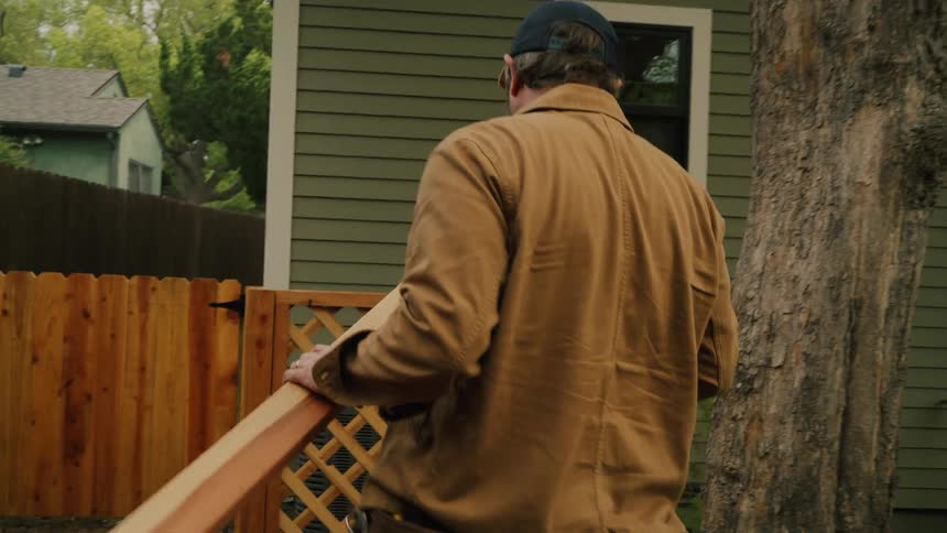 A man carries a long wooden plank he just cut across his backyard to install it on a new wooden fence