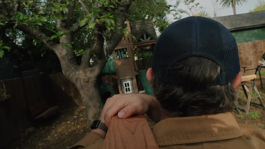 A man wearing a hat carries a heavy wooden beam on his shoulder as he walks through his backyard for a DIY construction project