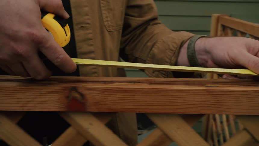 A person in a brown jacket measures a wooden lattice fence beam with a yellow tape measure in a backyard