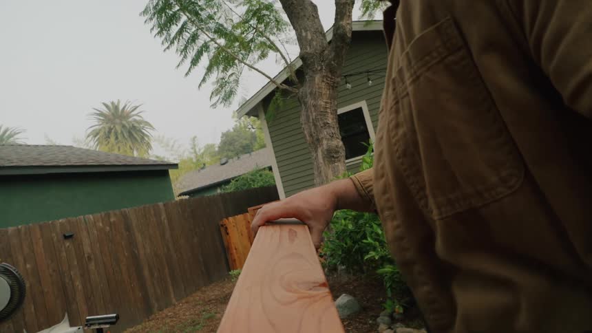 Cinematic slow-motion shot of a man's hands holding a piece of wood steady as a miter saw is lowered to make a cut in his backyard