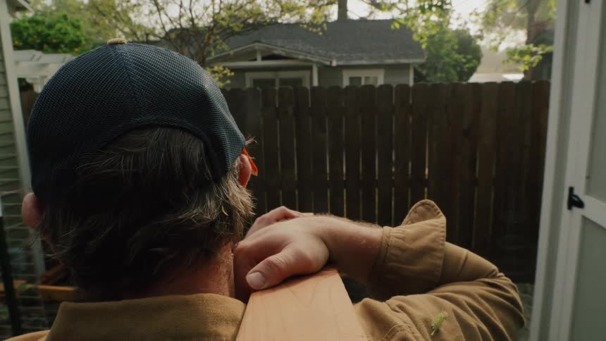 Over-the-shoulder slow-motion shot of a man carrying a large wooden beam through a backyard for a construction project