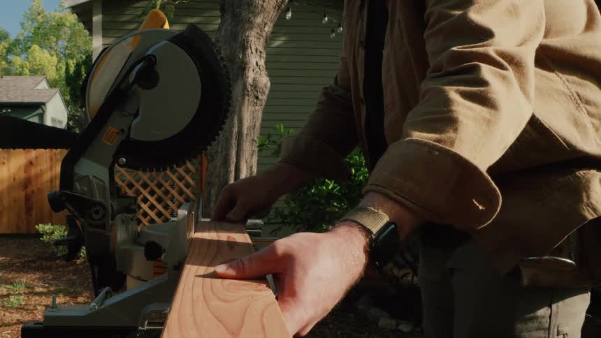A man in a work jacket carefully aligns a wooden plank on a miter saw for a precise cut in a sunny backyard