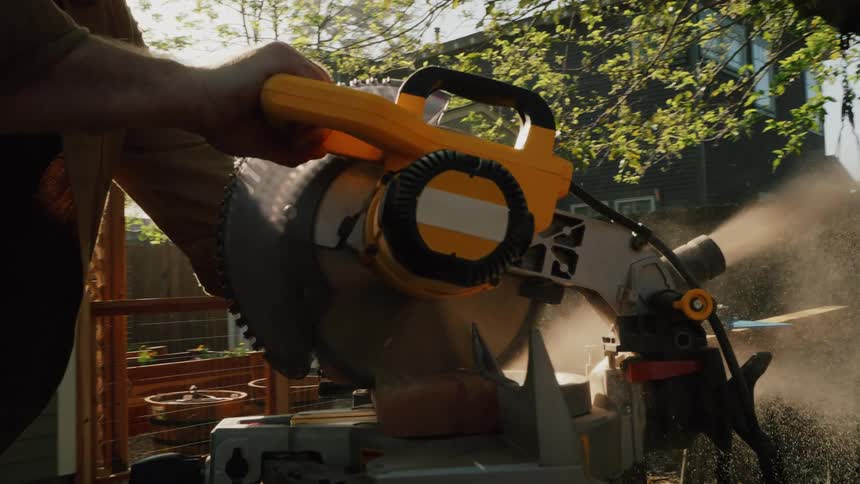 A man in his sunny backyard uses a miter saw to cut a piece of lumber in slow motion, sending sawdust flying