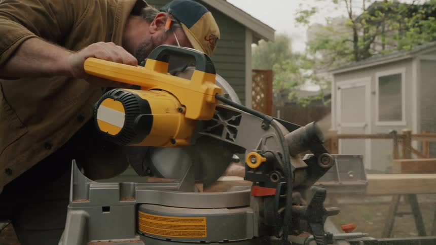 A carpenter carefully uses a miter saw to cut a piece of lumber in slow motion, with sawdust flying everywhere