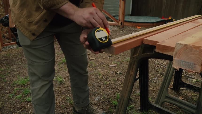 A man in a hat pulls a pencil from behind his ear to measure and mark a wooden plank on a sawhorse in his backyard