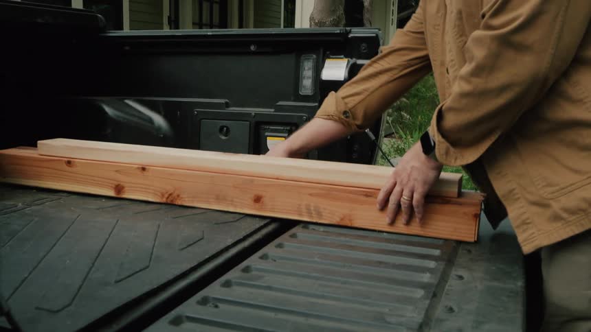 A carpenter wearing a tool belt unloads lumber from the bed of his pickup truck in a residential driveway