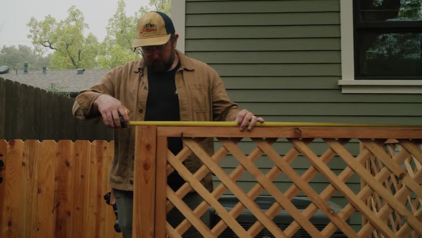 A man with a beard and hat carefully measures a new wooden lattice fence panel in a residential backyard