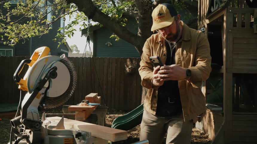A craftsman takes a break from a woodworking project to use his smartphone in a sunny backyard next to a miter saw