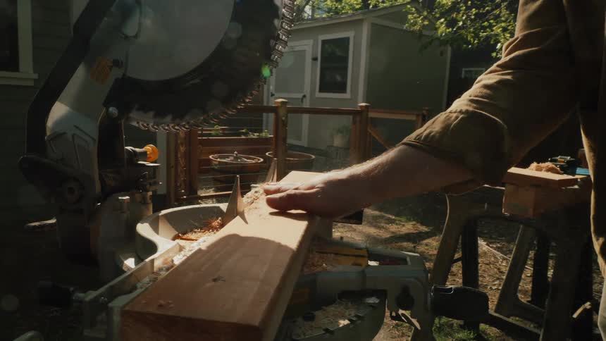 A woodworker clears sawdust from a wooden plank on a miter saw in a sunny backyard in slow motion