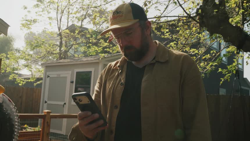 A carpenter wearing a hat and glasses scrolls through his phone in a sunlit backyard next to his miter saw