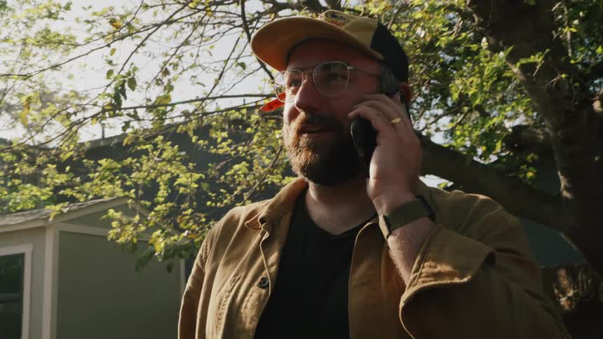 A bearded man wearing a baseball cap and glasses has a pleasant conversation on his smartphone in a sunny backyard