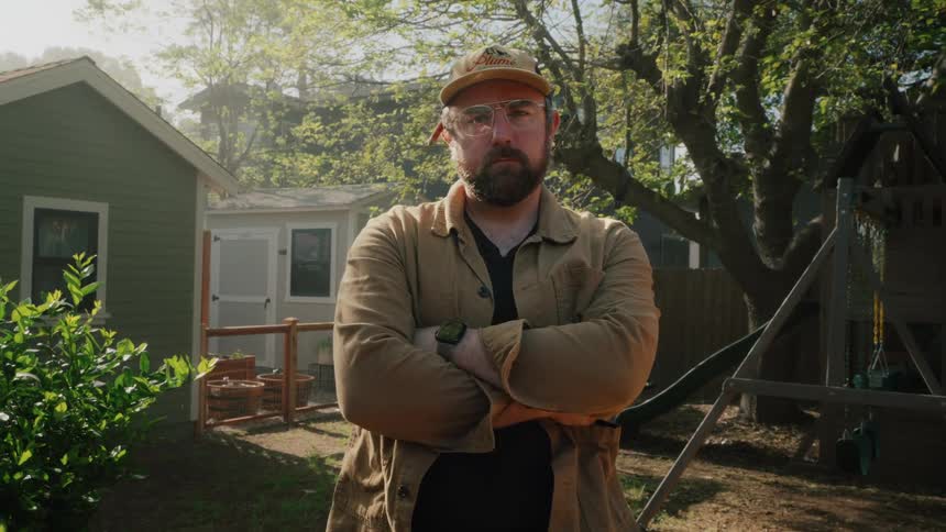 A serious handyman with a beard and glasses crosses his arms while looking at the camera in a sunny backyard