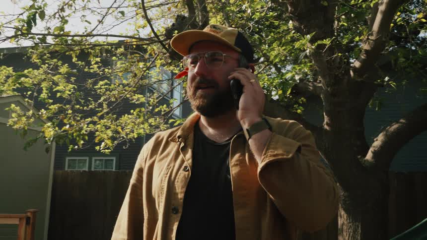 A smiling carpenter in a baseball cap and glasses takes a break to talk on the phone in his sunny backyard workspace