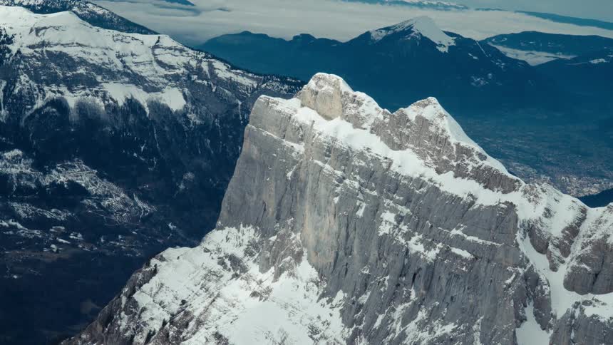 A cinemagraph captures the static, rugged peak of a snow-covered mountain towering above a vast valley and distant ranges
