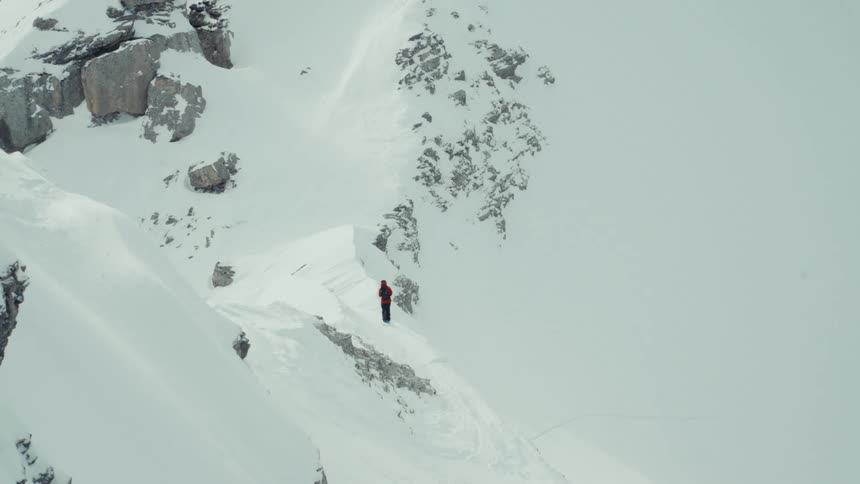 A lone skier in a red jacket stands on a remote, snow-covered mountain ridge