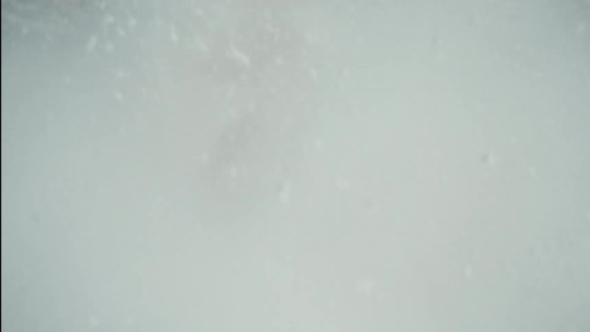 A skier's helmet is briefly seen as they carve hard, sending a huge cloud of powder snow directly into the camera in slow motion