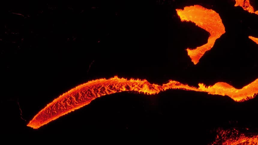 A high-angle shot captures a slow-moving river of molten lava glowing brightly against the dark, cooled volcanic rock
