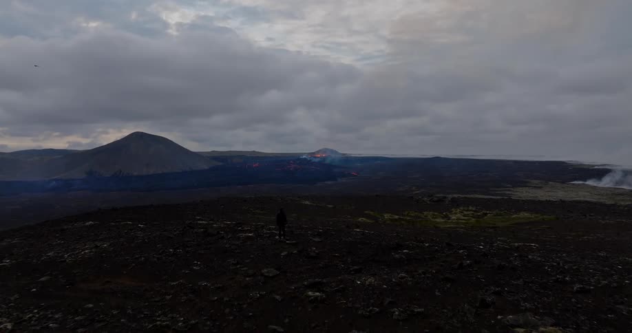 A lone figure stands in a vast, dark volcanic landscape watching the dramatic eruption of a distant volcano in Iceland