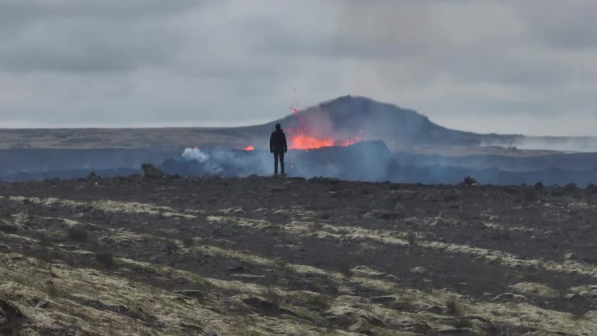 A lone adventurer stands silhouetted against a dramatic volcanic eruption, observing the fiery lava and smoke in a vast, desolate landscape