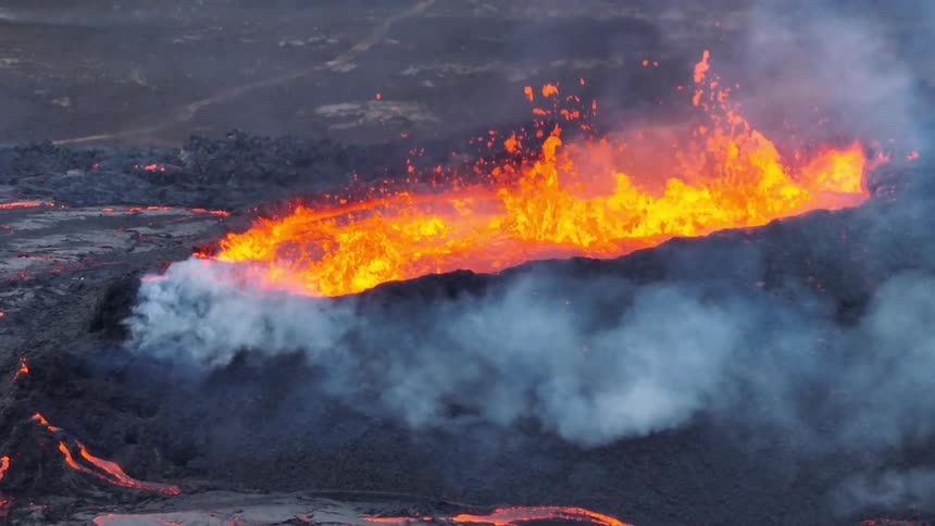 An epic aerial shot reveals a powerful volcanic eruption, with vibrant orange lava spewing from a fissure and flowing across the dark landscape
