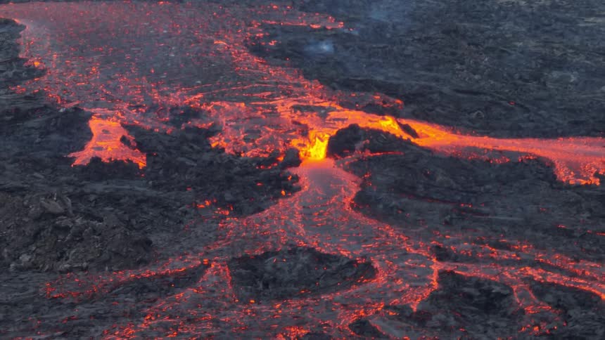 A mesmerizing wide shot captures a glowing river of molten lava flowing slowly over a vast field of dark, cooled volcanic rock