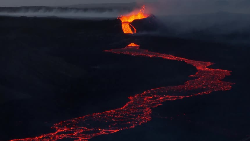 A wide shot reveals a river of glowing red lava flowing from an erupting volcano across a dark landscape at twilight