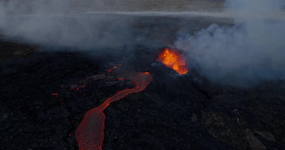 Static aerial view of an active volcano crater with a river of red hot lava flowing through dark, cooled rock under a smoky sky