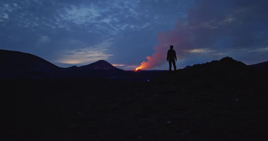 A lone silhouetted figure stands on a hill watching a dramatic volcanic eruption spewing lava and smoke at twilight