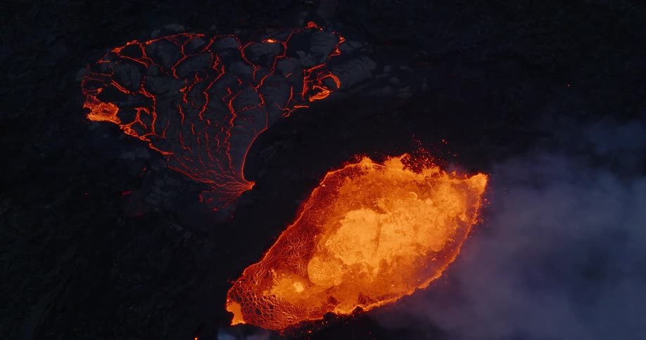 A dramatic aerial top-down view shows a bubbling caldera of molten lava beside a cooling, cracked lava field at night