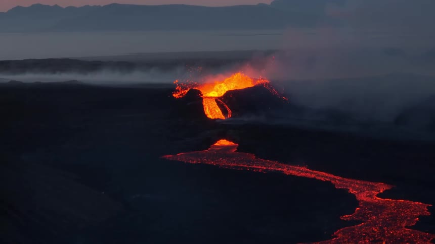 A dramatic wide aerial shot captures a volcano erupting at twilight, with glowing molten lava flowing down its slopes into a fiery river