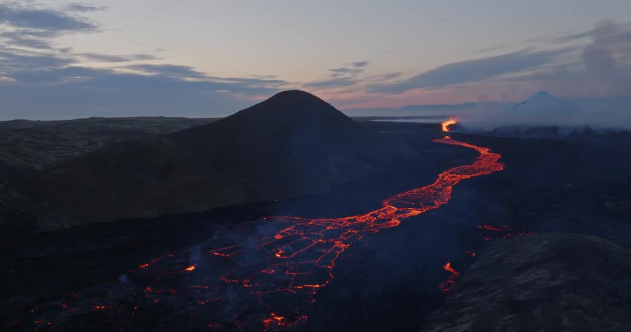 A breathtaking wide aerial shot shows a massive river of molten lava flowing from an erupting volcano at dusk