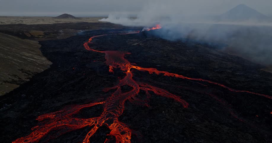 An epic aerial view pulls back from a glowing river of molten lava to reveal a vast, smoking volcanic eruption site