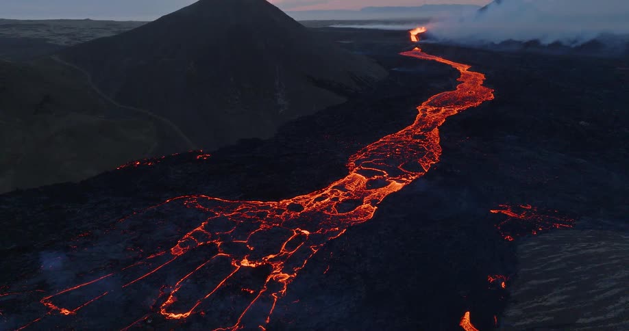 A breathtaking aerial shot reveals a glowing river of molten lava flowing from a distant erupting volcano through a vast, dark landscape at twilight