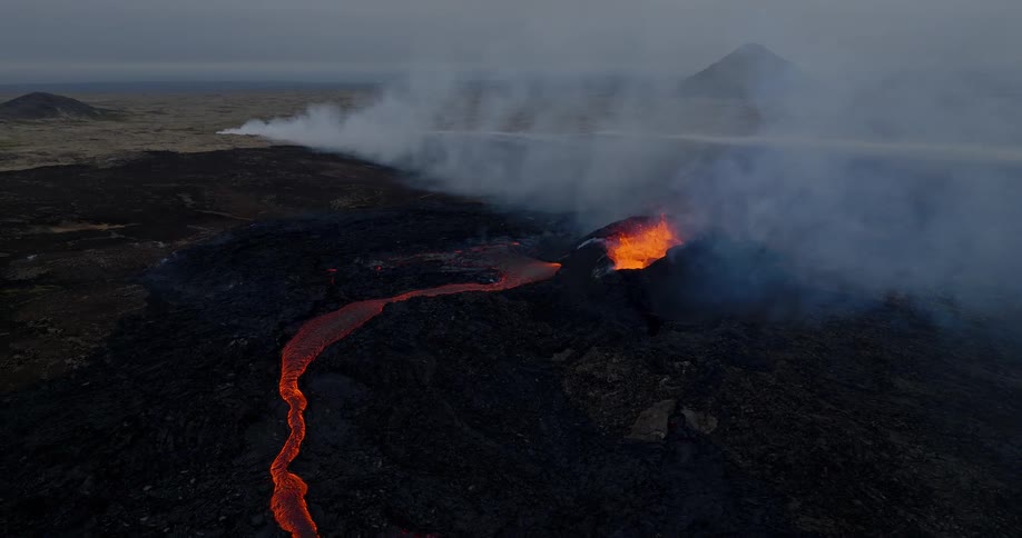 A dramatic aerial view captures a river of molten lava flowing from a volcanic crater, spewing smoke across a dark, barren landscape