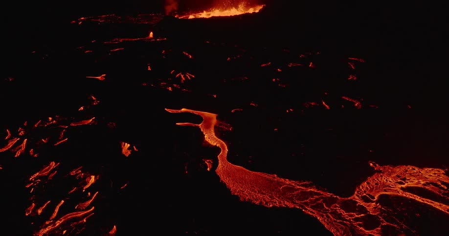 A dramatic aerial shot pulls back from a glowing river of molten rock to reveal a powerful volcanic fissure eruption at night