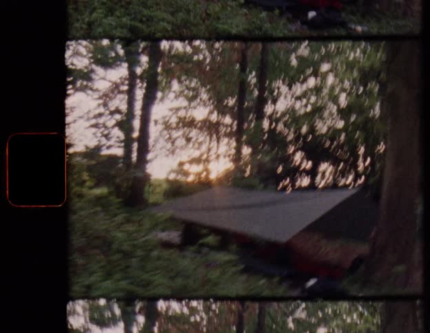 A shaky, vintage shot shows a young man emerging from behind a tree at a rustic campsite in the woods at golden hour