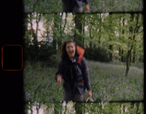 A woman with a backpack makes a funny face and smiles while hiking through a forest full of wildflowers in this vintage film footage